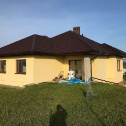 Żółty dom jednorodzinny z brązowym dachem z blachodachówki, widoczny trawnik, błękitny wąż ogrodowy, oraz fragment krajobrazu w tle.