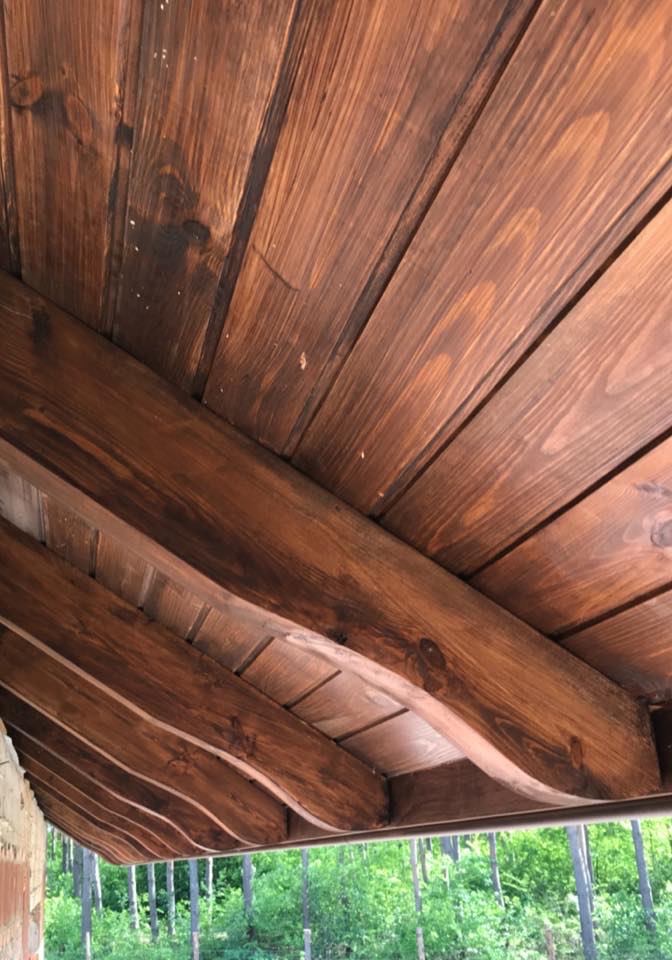 Drewniana podbitka dachowa z widocznymi belkami, pomalowana bejcą w odcieniach brązu, z widokiem na zieleń drzew w tle.