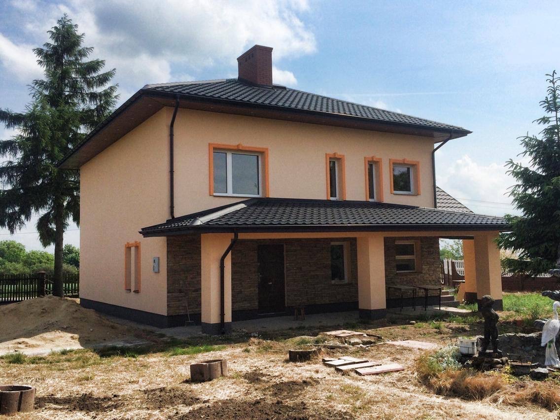 Dwukondygnacyjny dom murowany z ciemnym dachem, beżową elewacją z pomarańczowymi obramowaniami okien, częściowo wykończony kamieniem elewacyjnym na ganku, otoczony niezagospodarowanym terenem...