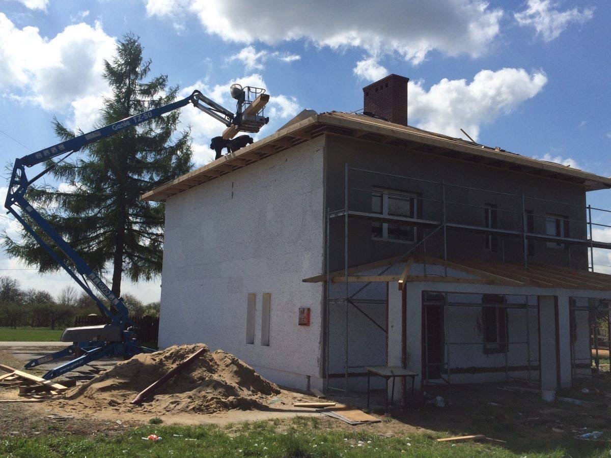 Dwukondygnacyjny dom w trakcie budowy z częściowo otynkowaną elewacją, rusztowaniami i podnośnikiem koszowym, na którym pracują dwie osoby przy przygotowaniu dachu, widoczny stos piasku.