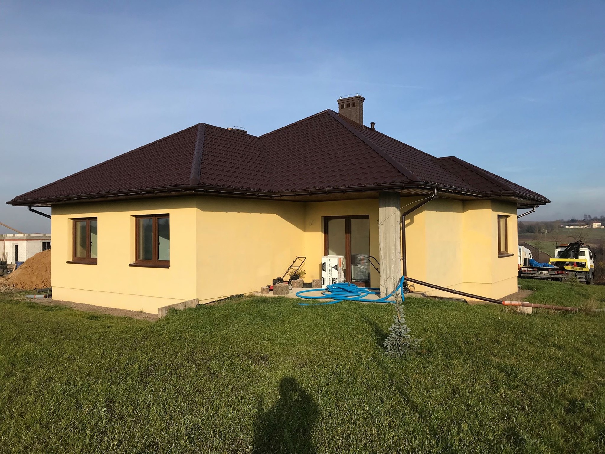 Żółty dom jednorodzinny z brązowym dachem z blachodachówki, widoczny trawnik, błękitny wąż ogrodowy, oraz fragment krajobrazu w tle.