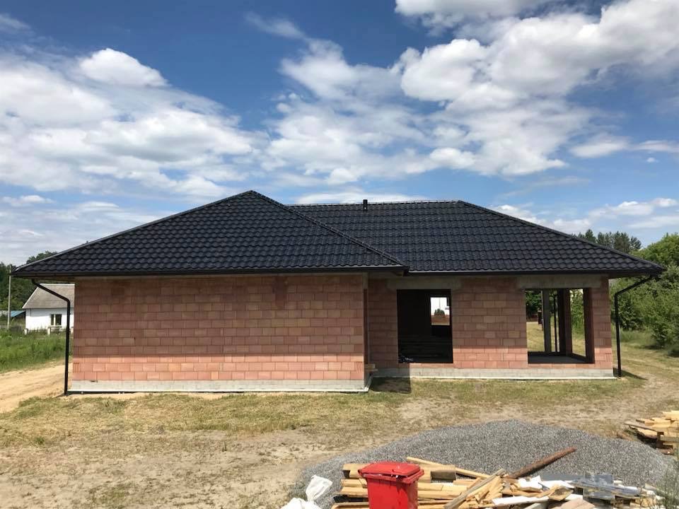 Niedokończony dom murowany z ciemnym dachem z blachodachówki, widoczne ściany z czerwonej cegły, otwory okienne i drzwiowe, rynna, w tle błękitne niebo z chmurami, na pierwszym planie gruz i drewno.