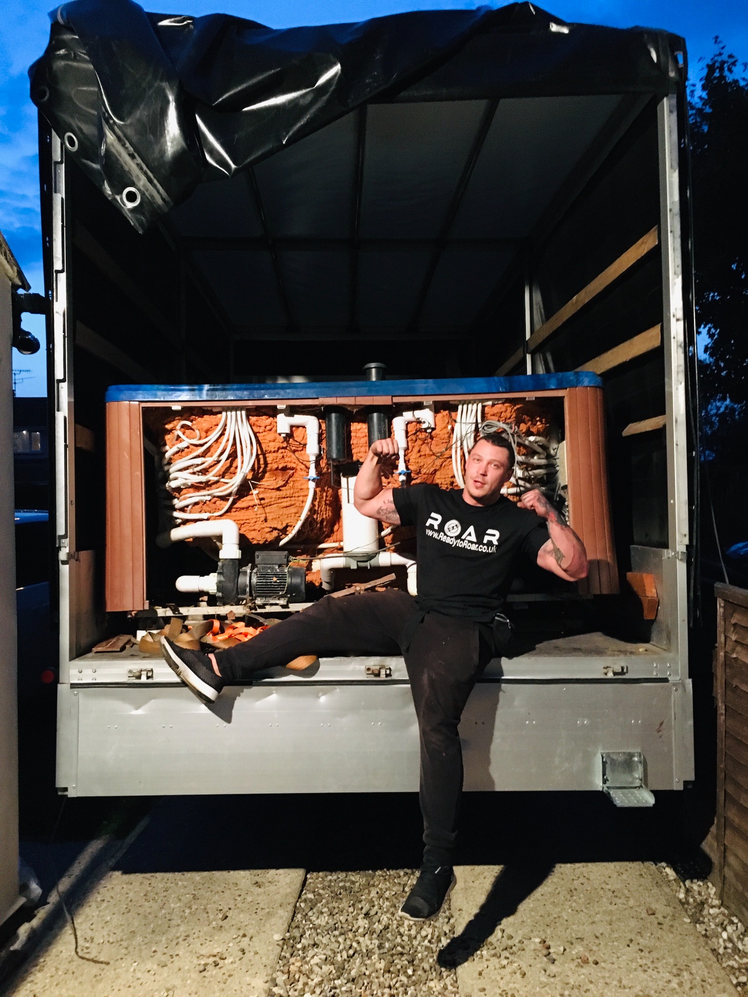 Mężczyzna w czarnej koszulce z logo, siedzący na rampie ciężarówki z odkrytą wanną z hydromasażem, eksponując jej wewnętrzne okablowanie i system filtracji.