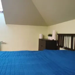 Poddasze z oknem dachowym, pomalowane na jasnożółty kolor, z widocznym fragmentem łóżka z niebieską narzutą, komodą i dekoracjami.