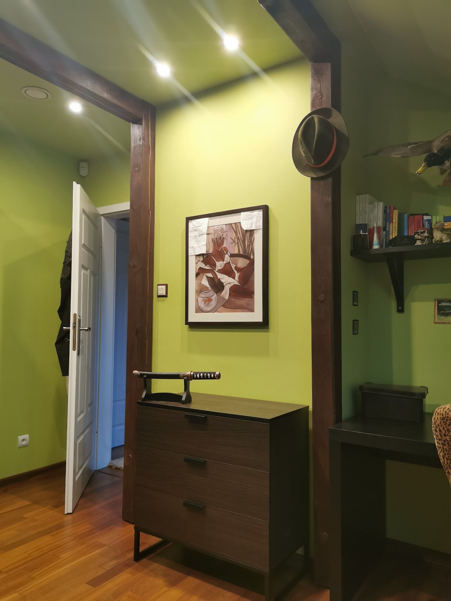 Jasnozielony pokój z drewnianymi belkami, komodą z mieczem samurajskim, obrazem na ścianie, otwartymi drzwiami i półką z książkami i trofeami myśliwskimi.