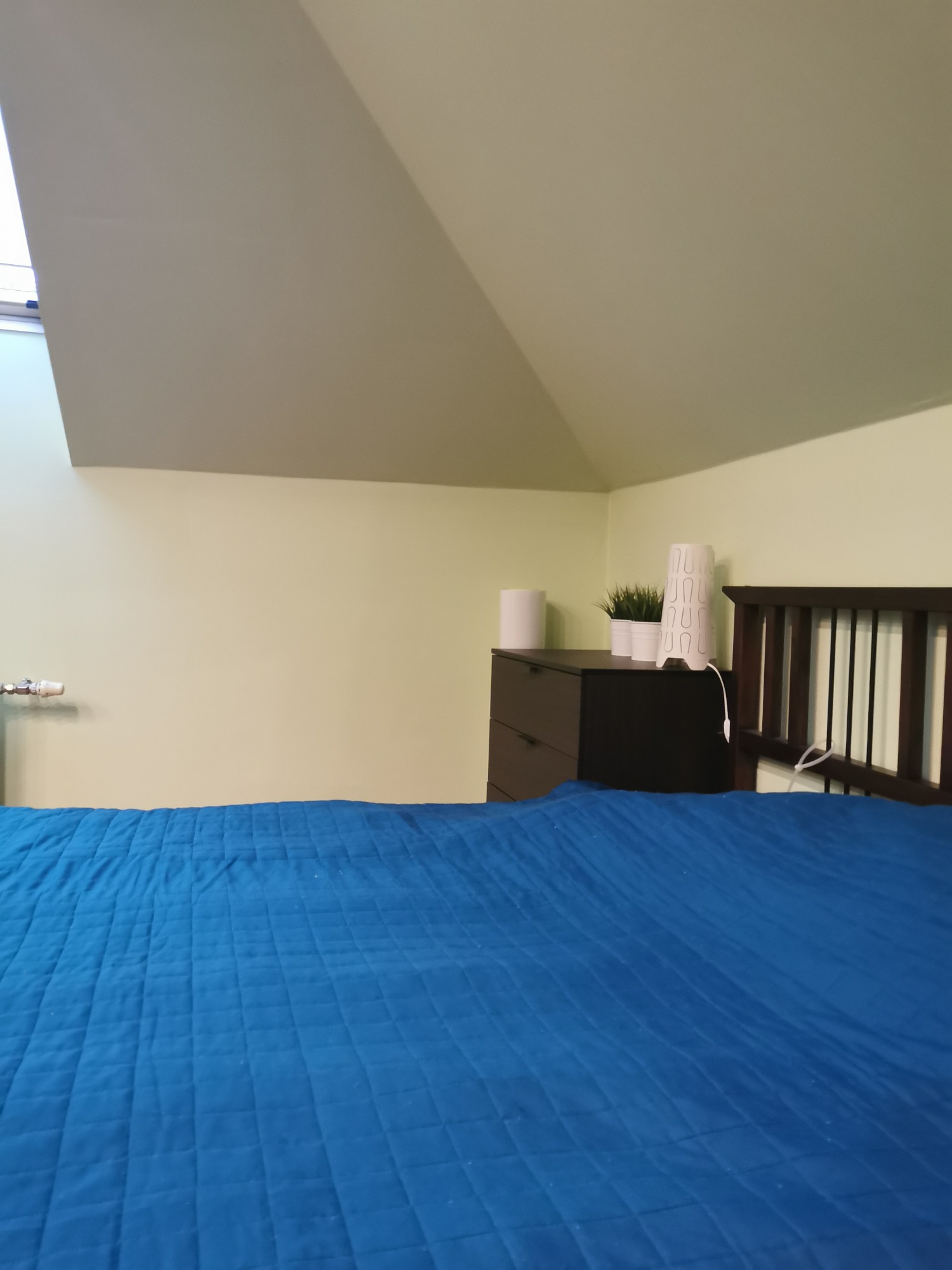 Poddasze z oknem dachowym, pomalowane na jasnożółty kolor, z widocznym fragmentem łóżka z niebieską narzutą, komodą i dekoracjami.