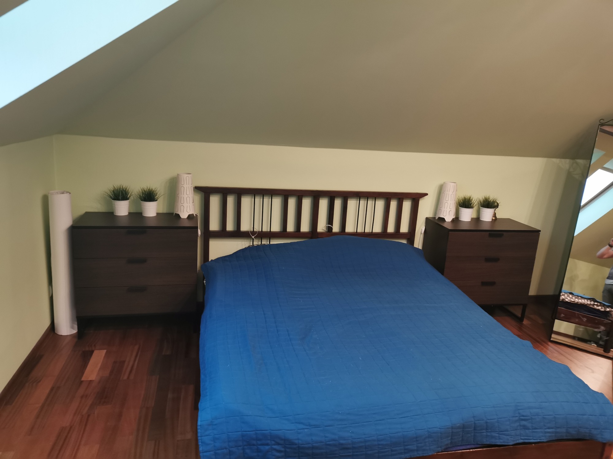 Pokój na poddaszu z drewnianą podłogą, łóżkiem z niebieską narzutą, dwiema szafkami nocnymi z dekoracjami i skośnym sufitem pomalowanym na jasnozielono.