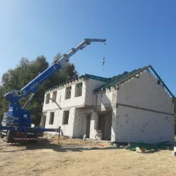 Wznoszenie dwukondygnacyjnego budynku z bloczków, widoczny dźwig budowlany Böcker z wysięgnikiem nad konstrukcją dachu, teren budowy z piaskiem i zielonymi elementami konstrukcyjnymi.