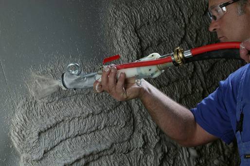 Aplikacja maszynowa tynku cementowo-wapiennego na ścianę, widoczna dysza pistoletu z czerwoną rurą i metalowymi elementami, operator w okularach ochronnych i niebieskim kombinezonie.