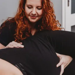 Fizjoterapeutka z rudymi włosami wykonuje masaż pleców pacjentki w czarnym ubraniu. Widoczny fragment białych drzwi i szarej ściany w tle.