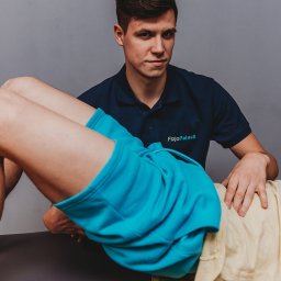 FizjoPaluch - nowoczesna fizjoterapia Sp. z o.o. - Młody mężczyzna w granatowej koszulce 'FizjoPaluch' wykonuje ćwiczenie z pacjentem leżącym na stole. Ujęcie z bliska, na szarym tle.