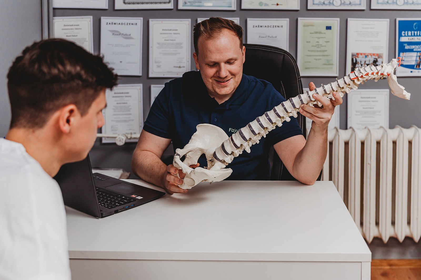 Lekarz prezentuje model kręgosłupa pacjentowi w gabinecie. Na ścianie widoczne certyfikaty. Ujęcie z boku, z bliska.