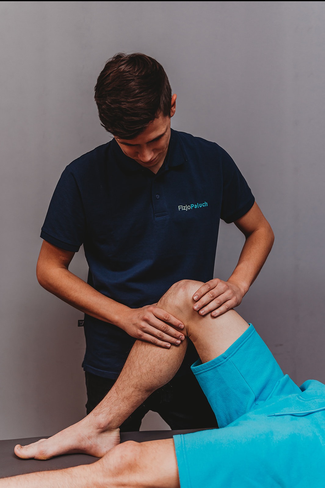 Fizjoterapeuta w granatowej koszulce z logo 'FizjoPaluch' masuje kolano pacjenta leżącego na leżance w jasnoniebieskim ubraniu, na szarym tle.