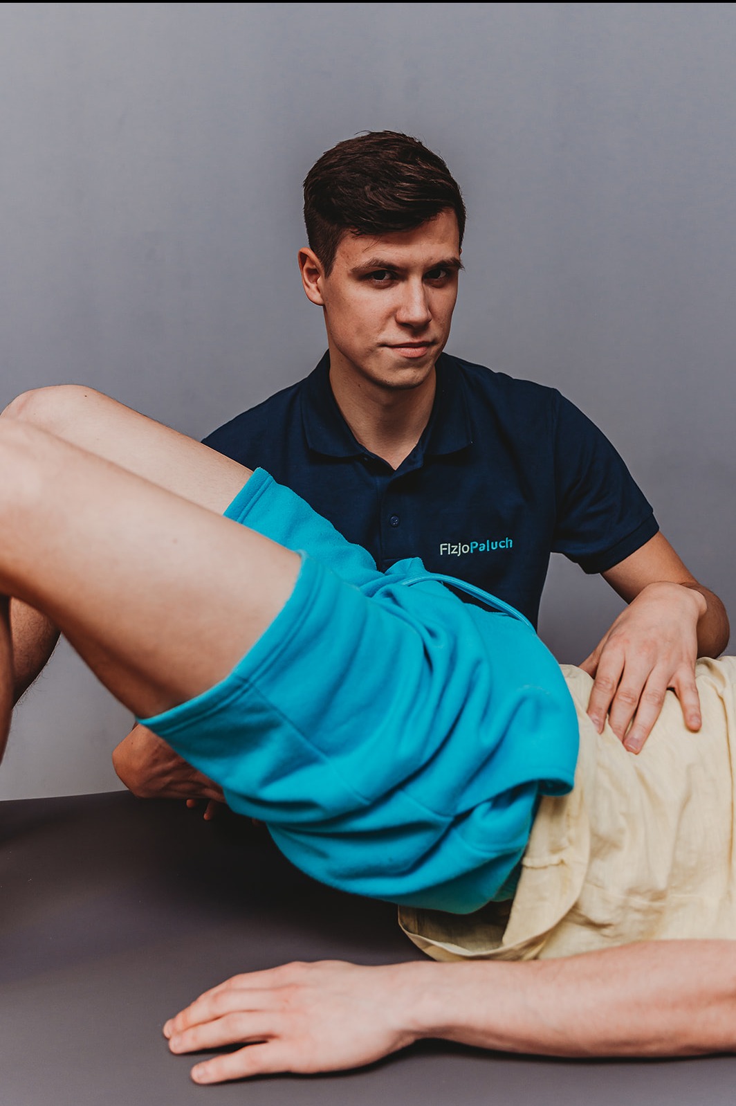 Młody mężczyzna w granatowej koszulce 'FizjoPaluch' wykonuje ćwiczenie z pacjentem leżącym na stole. Ujęcie z bliska, na szarym tle.