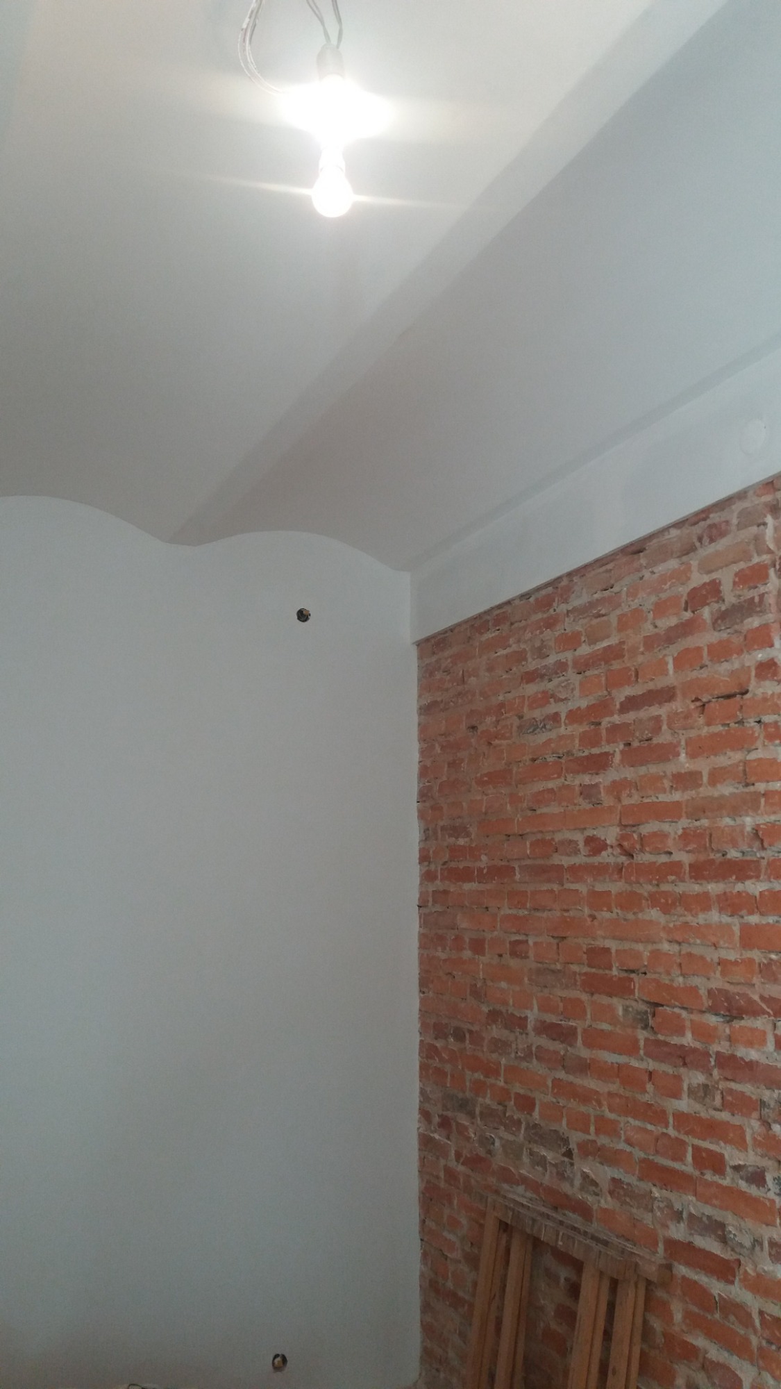 Świeżo pomalowane na biało ściany w pomieszczeniu z fragmentem odsłoniętej, ceglanej ściany i widoczną żarówką na suficie.