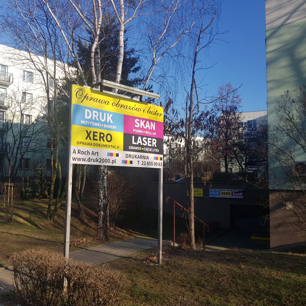 Zewnętrzny szyld reklamowy drukarni z ofertą: druk wizytówek i banerów, ksero, skan, laser - grawer i cięcie liter, oprawa obrazów i luster, pianki roll-up, oprawa dokumentacji. Widok na budynek...