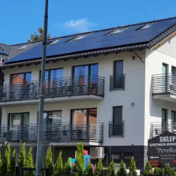 Trzypiętrowy budynek z białymi ścianami i balkonami, na dachu zainstalowane panele fotowoltaiczne. Widoczny szyld sklepu 'Perełka'.