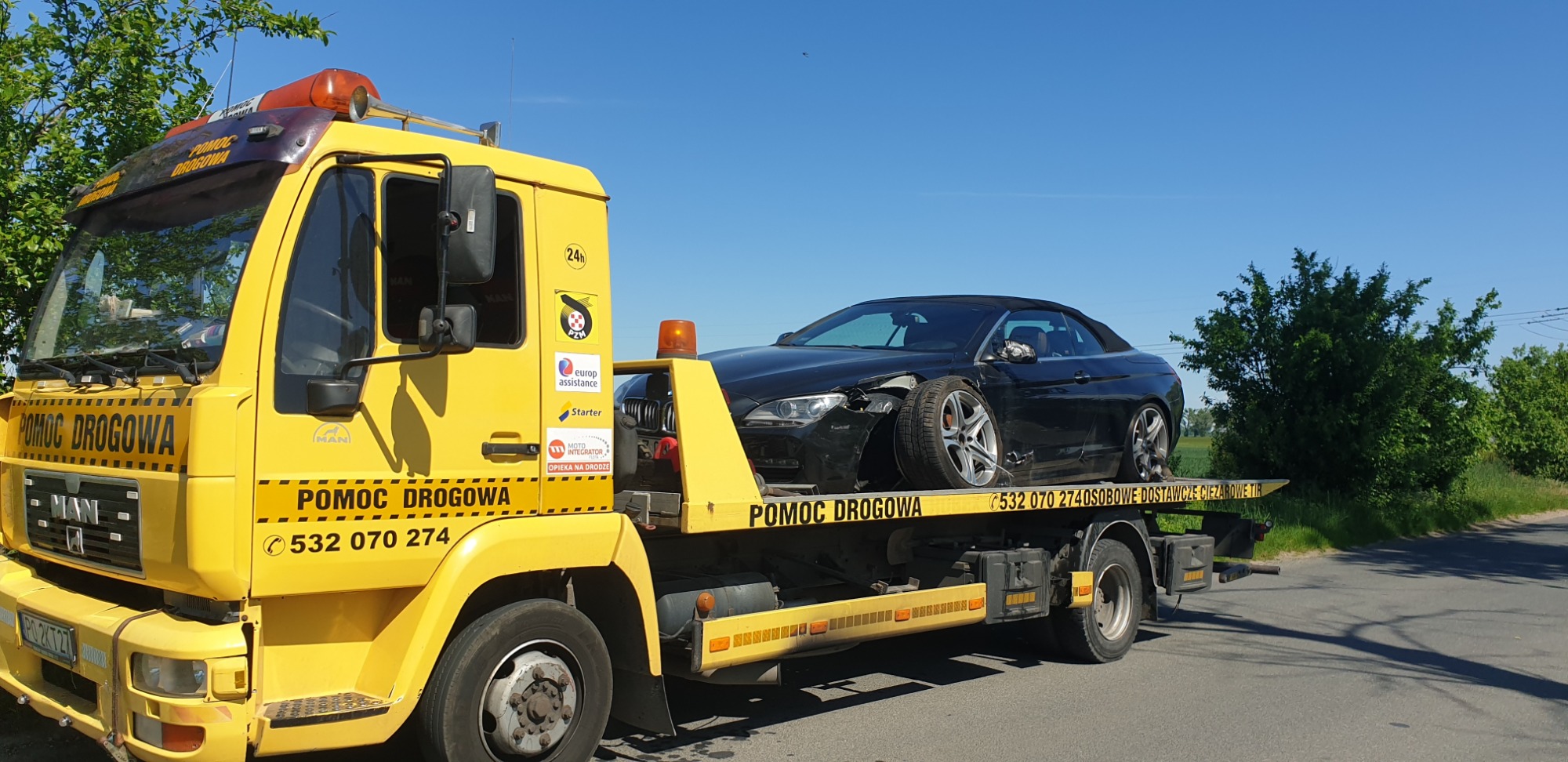 Żółta laweta z uszkodzonym czarnym kabrioletem BMW na platformie, widoczne logo firm i numer telefonu na boku pojazdu pomocy drogowej.