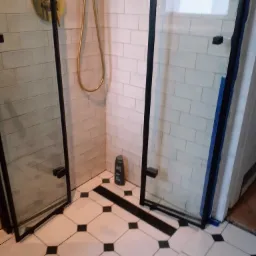 Kabina prysznicowa z czarnymi ramami i złotą armaturą na tle białych, cegiełkowych płytek i podłogi w biało-czarną szachownicę; butelka szamponu w rogu.