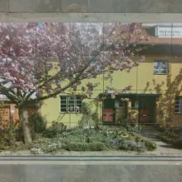 Mozaika ceramiczna przedstawiająca żółty dom z czerwonymi drzwiami, kwitnącym drzewem i ogrodem, ułożona na ścianie z szarych płytek.