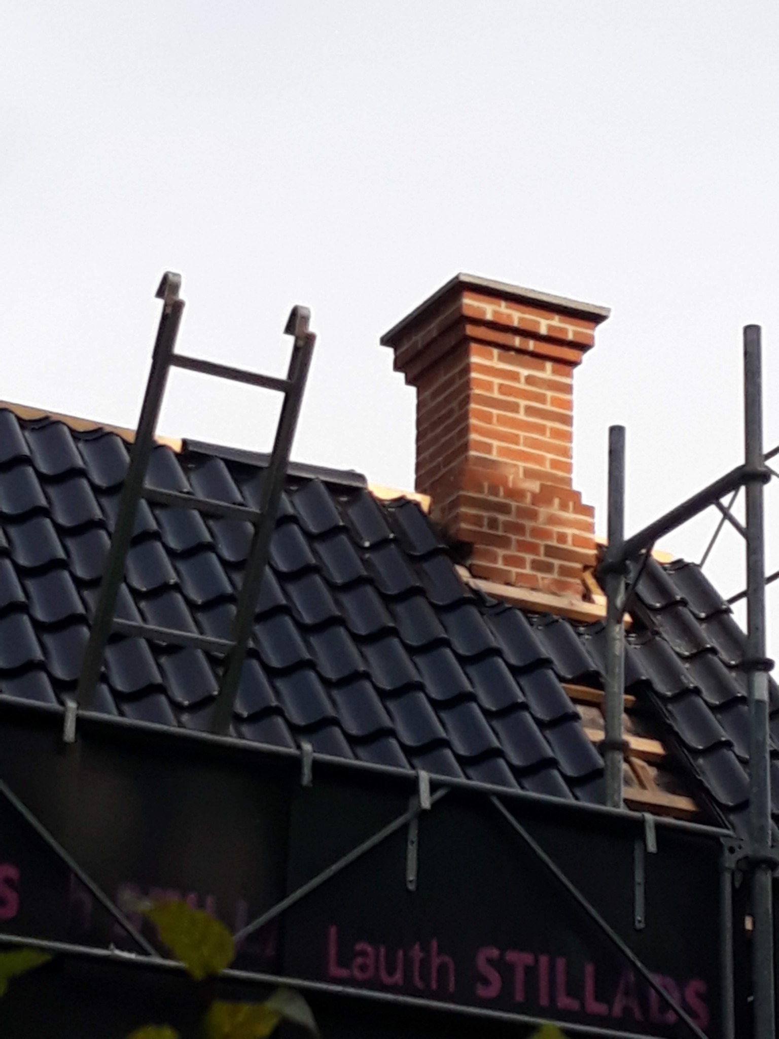 Ceglany komin na dachu z czarnej dachówki, częściowo zasłonięty rusztowaniem i drabiną; widoczny fragment baneru reklamowego z napisem.