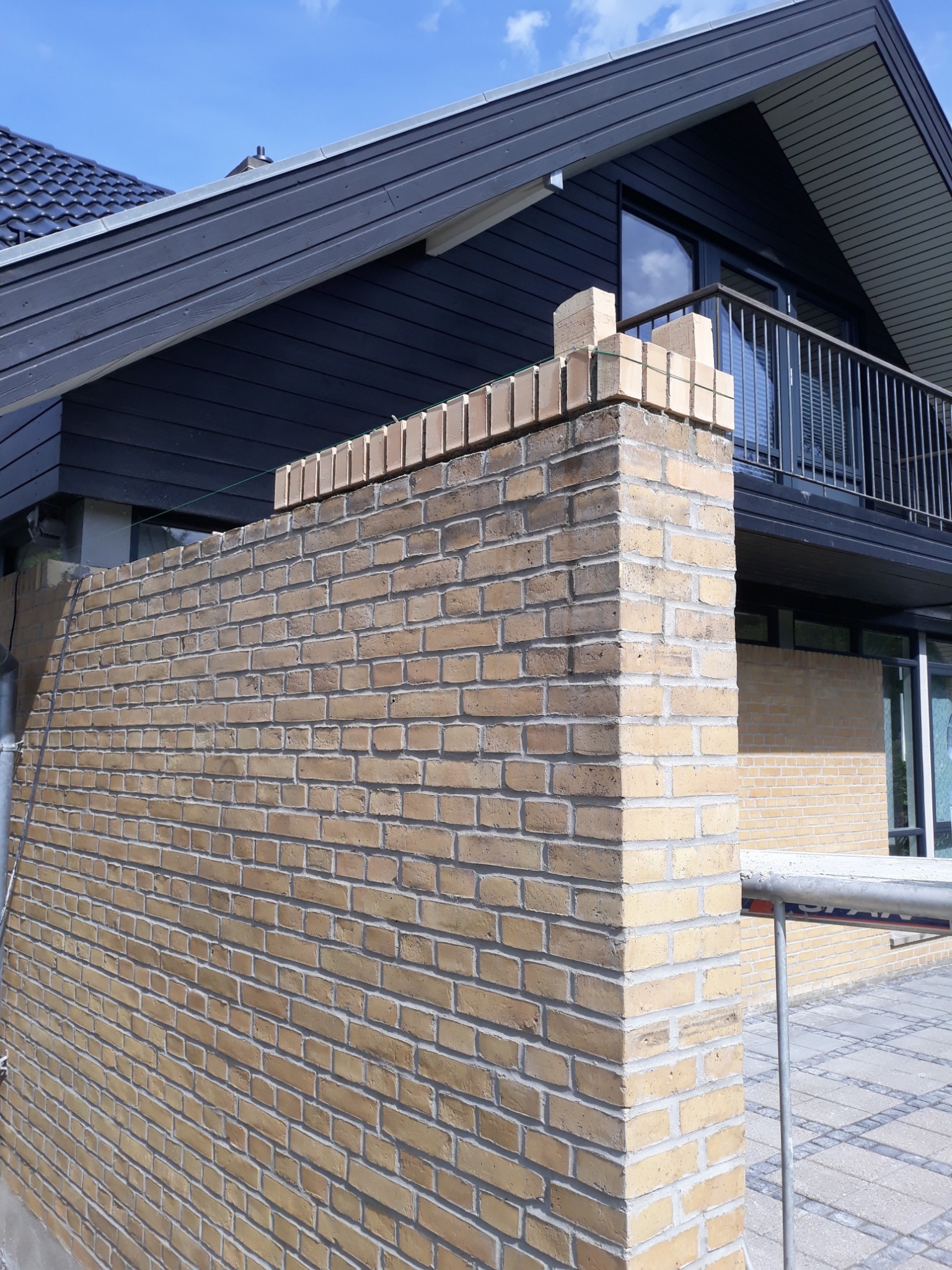 Wznoszenie muru z żółtej cegły klinkierowej na tle nowoczesnego domu z czarną elewacją i balkonem. Widoczne świeżo ułożone cegły na szczycie muru, zabezpieczone zielonym sznurkiem. Fragment...