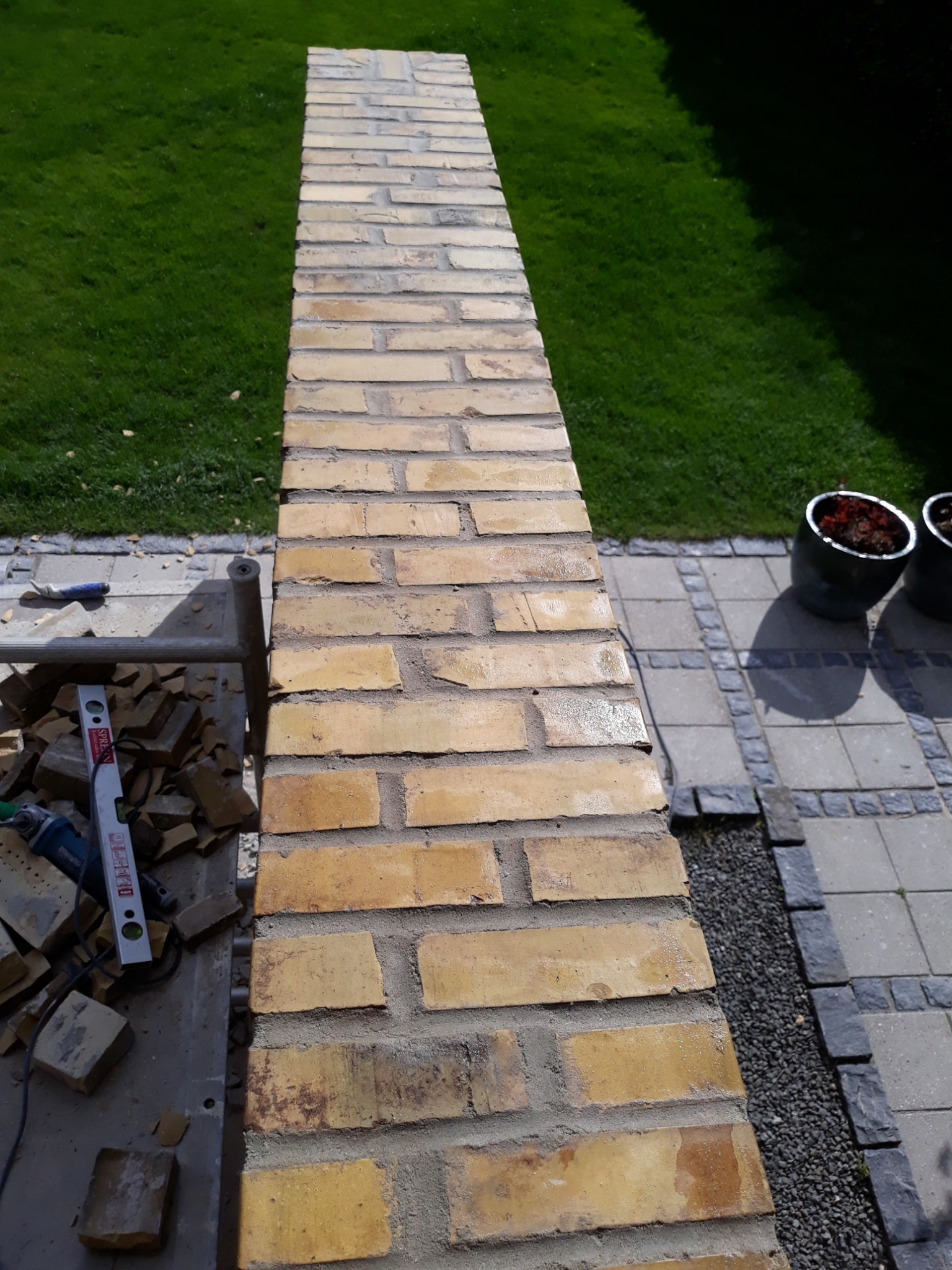 Wznoszenie murku z żółtej cegły klinkierowej w Lęborku, widoczne narzędzia murarskie, poziomica i fragment trawnika w tle.