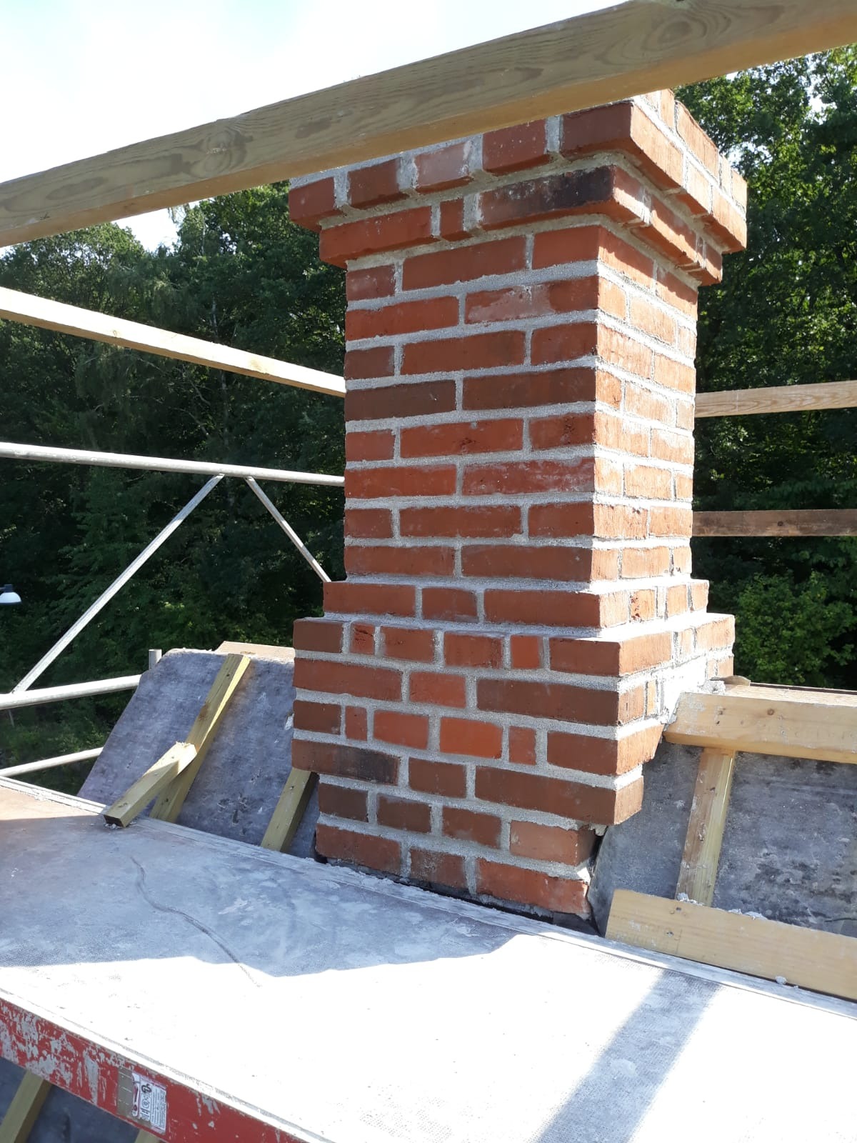 Murowany komin z czerwonej cegły, w trakcie budowy na dachu, widoczne rusztowanie i elementy konstrukcyjne dachu.