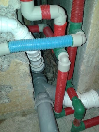 Złożona instalacja wodno-kanalizacyjna z rur PCV w kolorach: białym, szarym, czerwonym, zielonym i niebieskim, widoczna w wykuciu w starej ścianie.