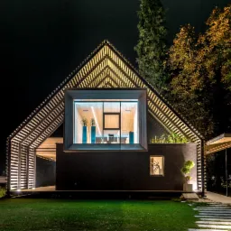 Nowoczesny dom z drewnianą konstrukcją szkieletową podświetloną nocą, widoczne duże okno z roślinami wewnątrz.