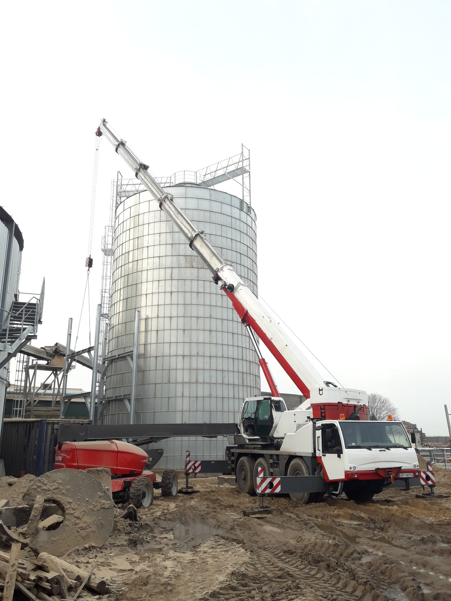 Montaż srebrnego, cylindrycznego zbiornika przemysłowego z użyciem białego dźwigu samochodowego z czerwonymi elementami, widoczny również czerwony podnośnik teleskopowy Manitou na błotnistym placu...