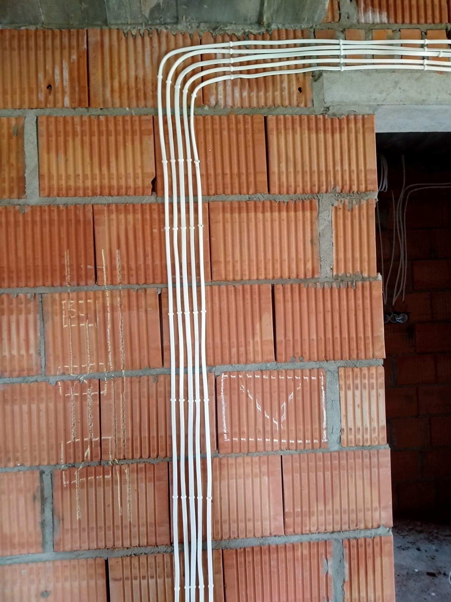 Białe przewody elektryczne prowadzone wzdłuż ściany z czerwonej cegły dziurawki, zamocowane za pomocą uchwytów. Na cegłach widoczne białe oznaczenia rysunkowe, prawdopodobnie schemat instalacji.