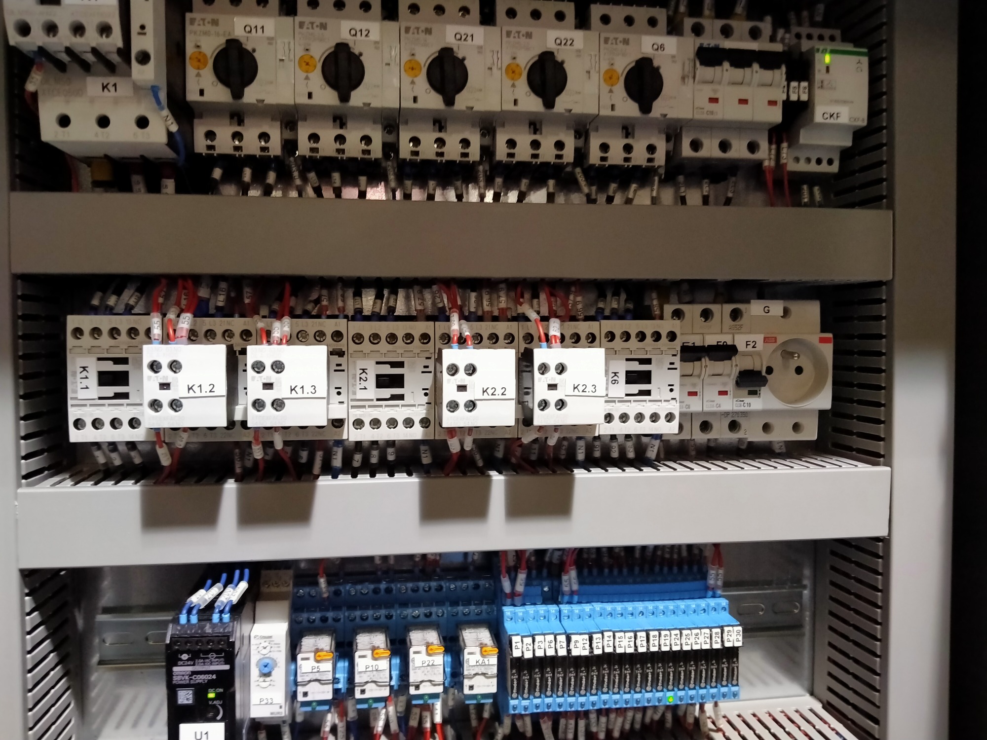 Rozdzielnica elektryczna z widocznymi bezpiecznikami, stycznikami i okablowaniem, w szarej obudowie, z elementami oznaczonymi etykietami.