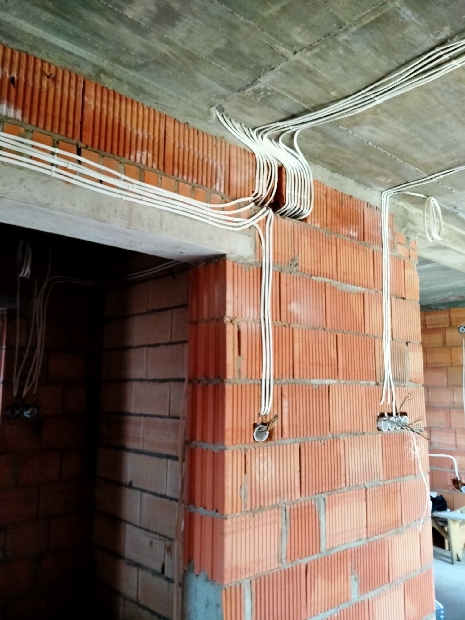 Wnętrze w trakcie budowy z widocznymi przewodami elektrycznymi prowadzonymi po ścianach z cegły i pod betonowym sufitem. Przewody zgrupowane przy otworach na puszki elektryczne.