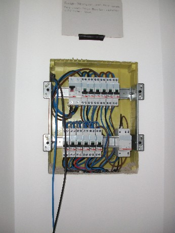 Otwarta skrzynka elektryczna z zainstalowanymi bezpiecznikami i przewodami w kolorach niebieskim, brązowym i czarnym, zamontowana na białej ścianie, nad skrzynką przyklejona kartka z zapiskami.