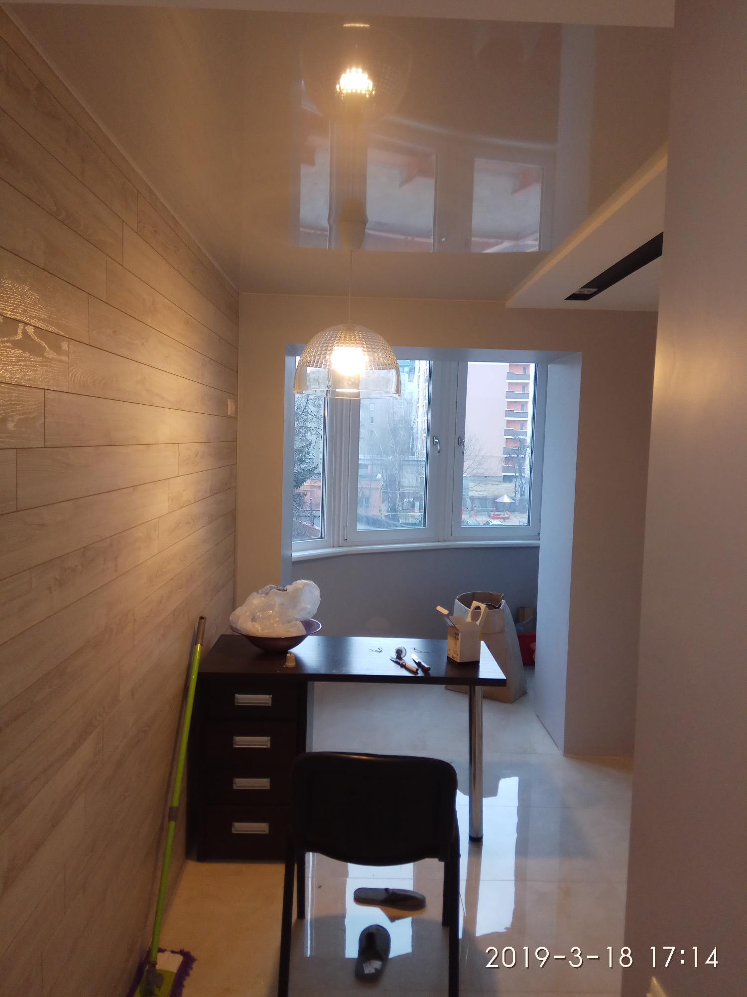 Wnętrze pokoju po remoncie z nowym oświetleniem: lampa wisząca z kloszem nad stołem, błyszczący sufit z odbiciem światła, ściana wyłożona panelami imitującymi drewno, widok na osiedle przez okno.