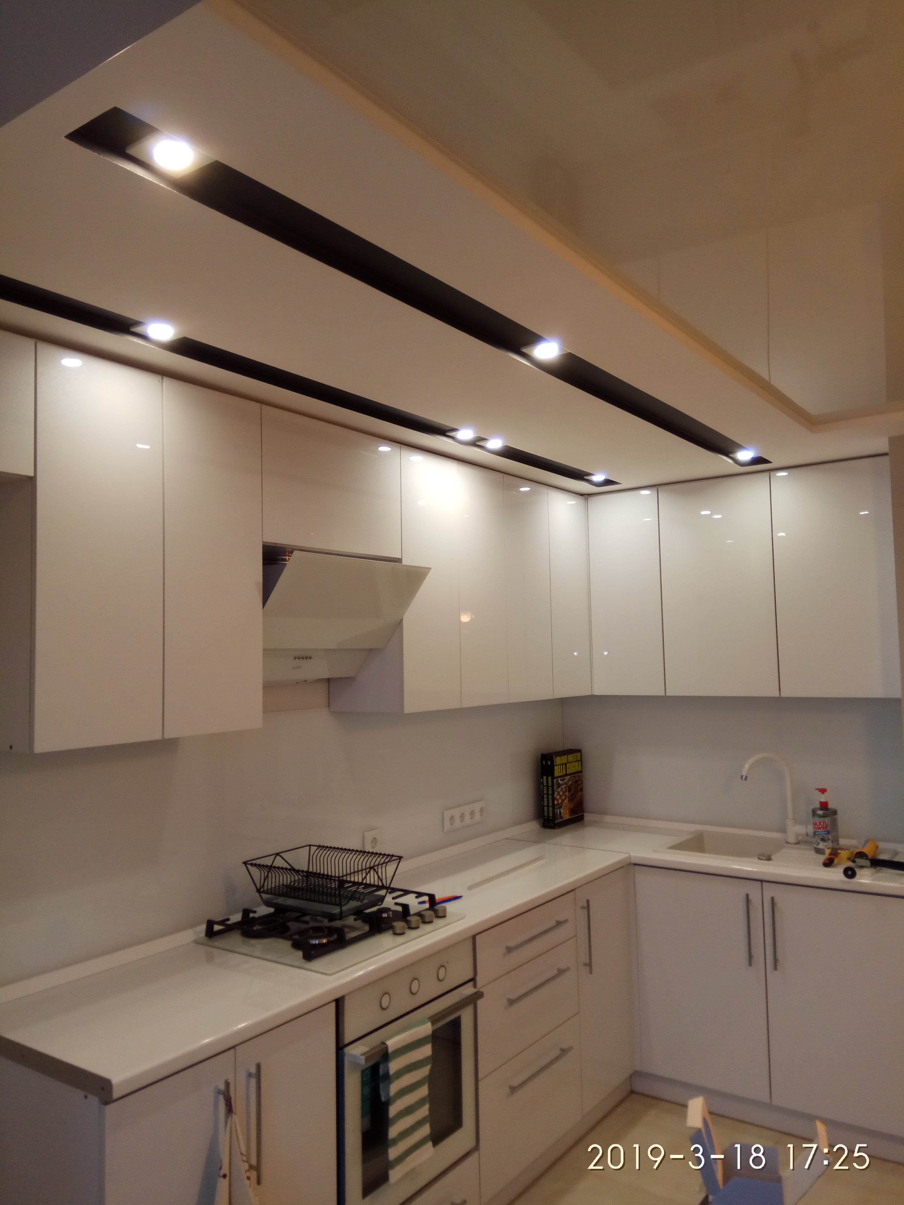 Nowoczesna, biała kuchnia z błyszczącymi frontami, oświetleniem punktowym w podsufitce i płytą gazową z suszarką na naczynia. Widoczne gniazdka elektryczne na ścianie.