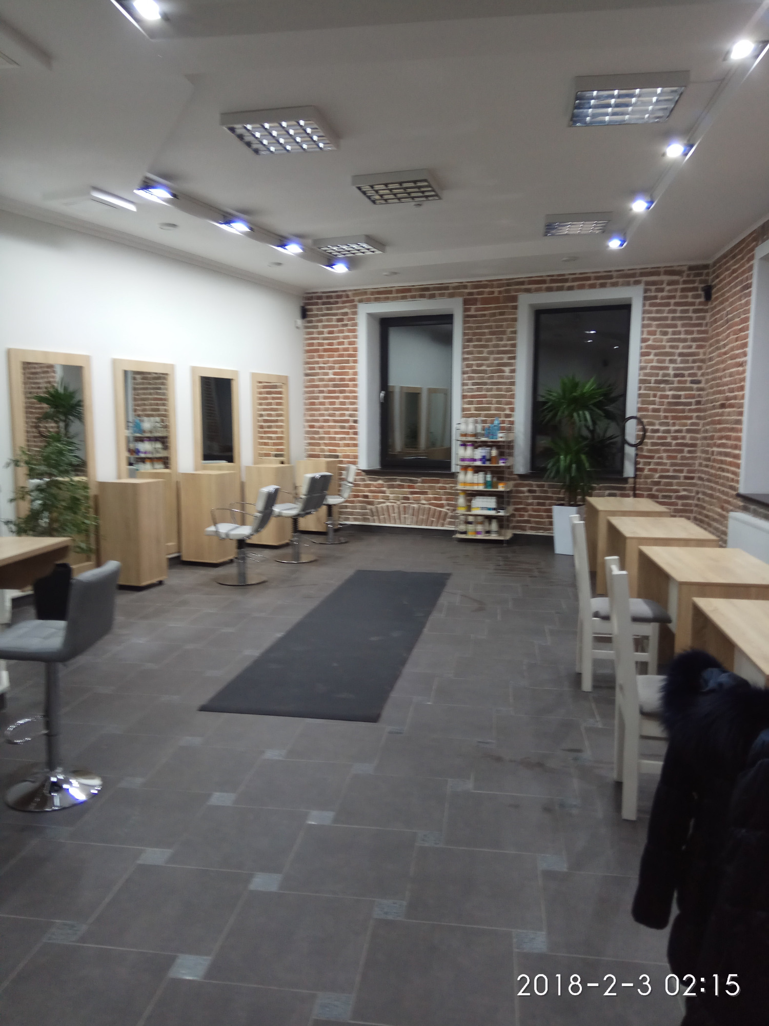 Jasne wnętrze salonu fryzjerskiego z czterema stanowiskami, każde z lustrem i krzesłem, oraz stanowiskami do manicure z białymi krzesłami, oświetlone panelami LED na suficie i lampami punktowymi.