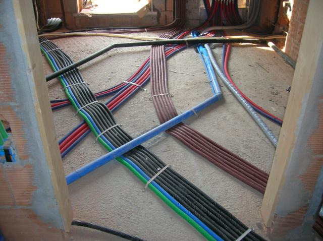 Wiązki przewodów elektrycznych w peszlach o różnych kolorach (czerwony, niebieski, zielony) ułożone na surowej podłodze w trakcie instalacji w nowym budynku, widoczne fragmenty muru z cegieł.