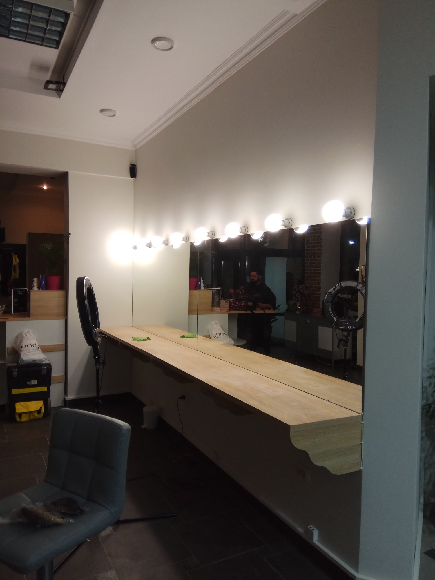 Długa toaletka z lustrem i rzędem żarówek wizażysty, oświetlenie sufitowe, ring lamp stojący obok, odbicie osoby w lustrze.