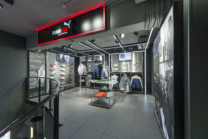 Wnętrze sklepu Puma Lab z oświetleniem punktowym, neonowym napisem i ekspozycją odzieży oraz obuwia sportowego na metalowych półkach i wieszakach.