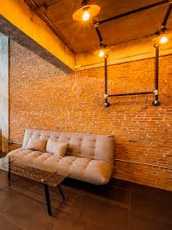 Loftowe wnętrze z ceglaną ścianą oświetloną trzema lampami z żarówkami Edisona, z widocznymi rurami instalacji elektrycznej. Pod ścianą stoi beżowa sofa i stolik kawowy.