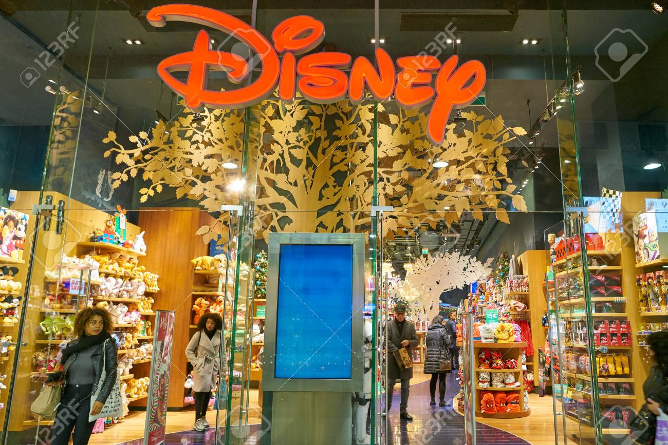 Wejście do sklepu Disney z pomarańczowym logo na szklanej fasadzie i dekoracyjnym drzewem w tle, widoczne półki z zabawkami i klienci.