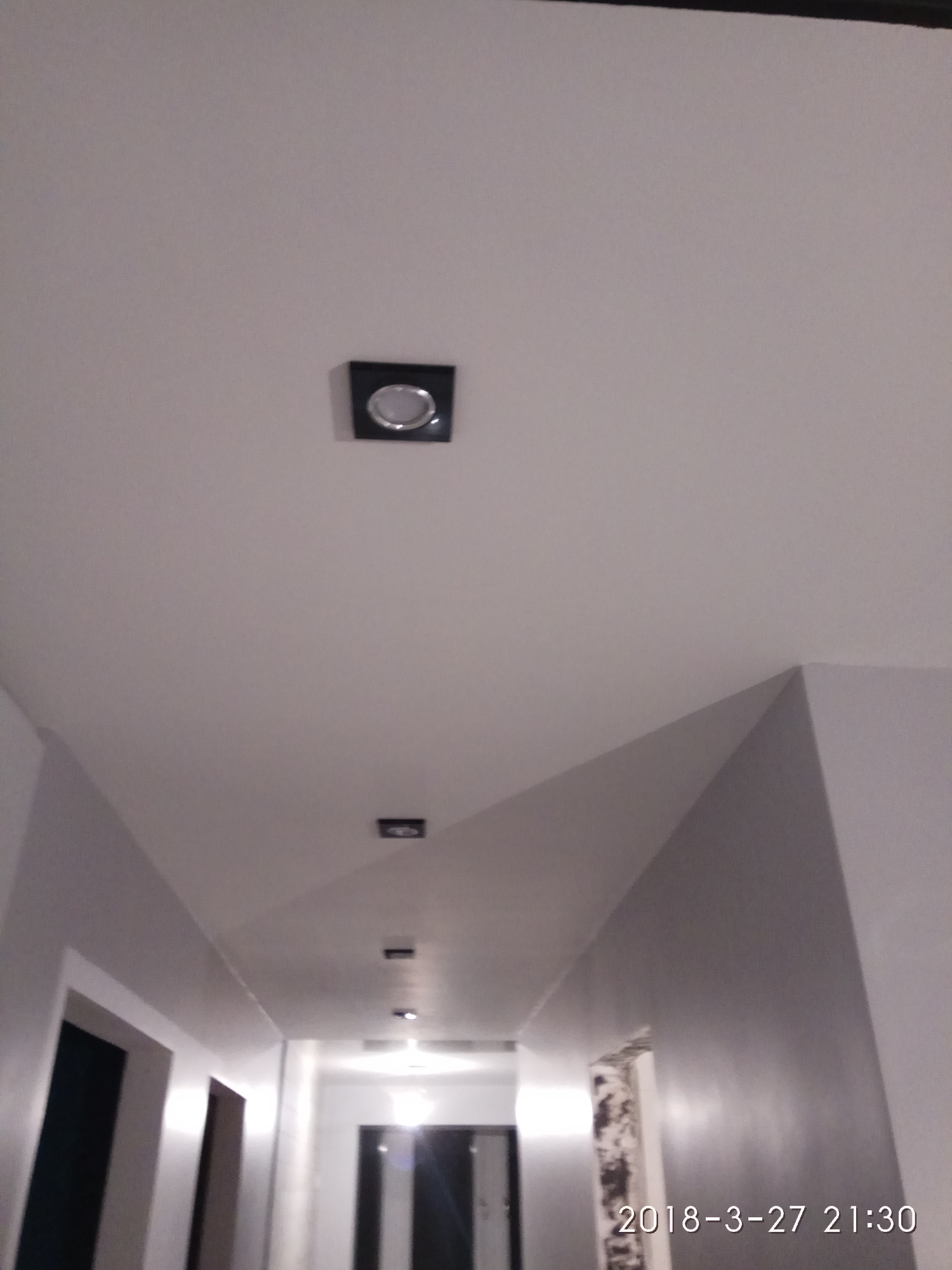 Korytarz z zamontowanymi kwadratowymi oprawami oświetleniowymi w suficie podwieszanym, widok od dołu, ciepłe światło.