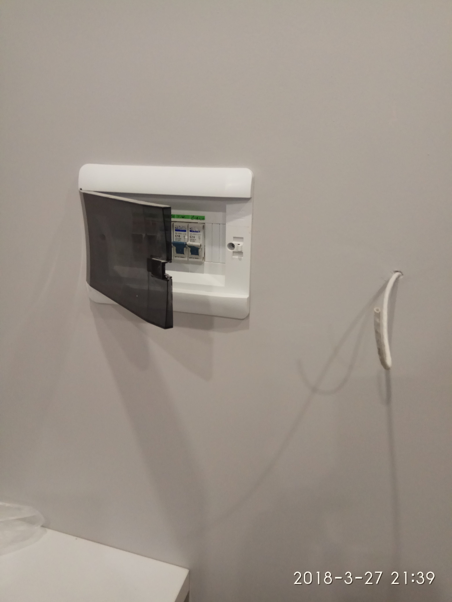 Skrzynka bezpiecznikowa z uchyloną, dymioną pokrywą na szarej ścianie. Obok widoczny wystający kabel elektryczny. Data i godzina wykonania zdjęcia widoczne w prawym dolnym rogu.