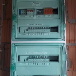 Rozdzielnica elektryczna z bezpiecznikami, opisana etykietami 'Zasilanie', 'Zabezpieczenia parter', 'Zabezpieczenia góra i piwnica', zamontowana na ścianie z cegły, z widocznymi przewodami w górnej...