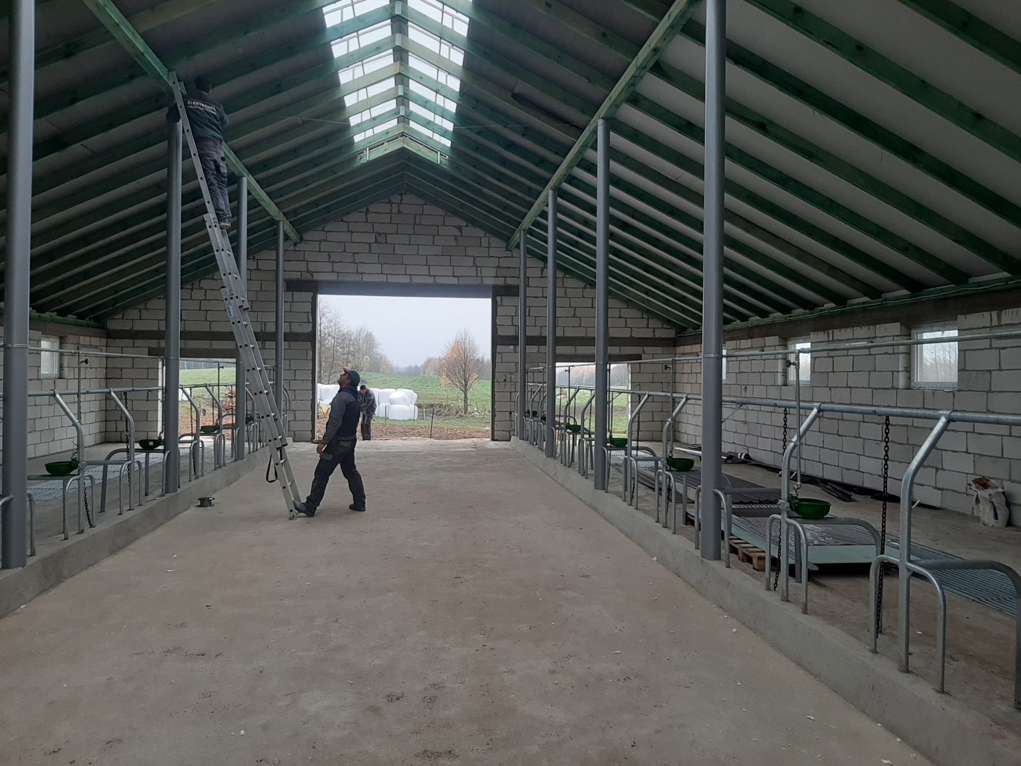 Wnętrze budowanego budynku gospodarczego z widocznymi elementami konstrukcyjnymi dachu i ścian z białego bloczka, pracownik na drabinie przy dachu, drugi pracownik na posadzce.