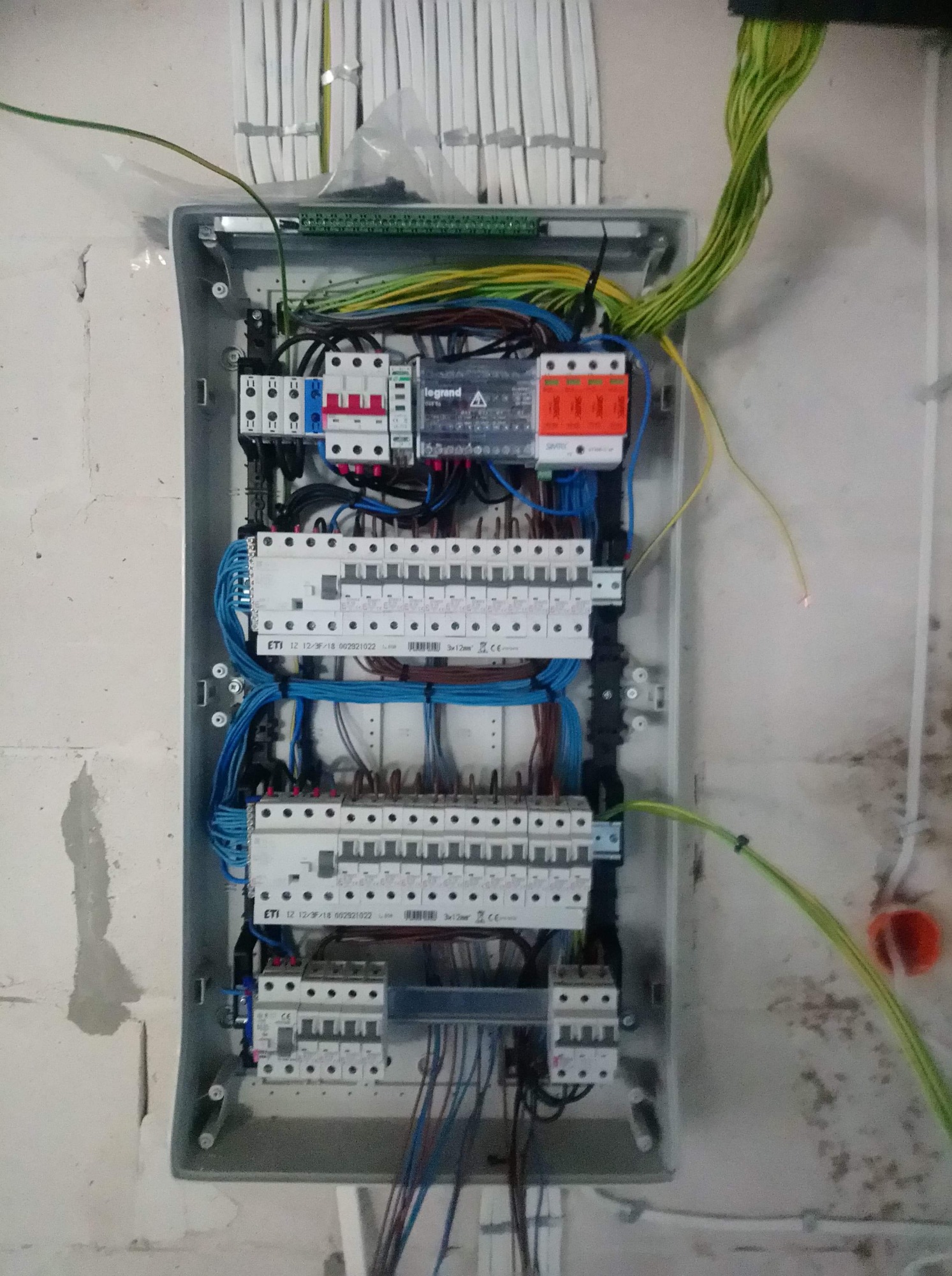 Otwarta szara skrzynka elektryczna z zainstalowanymi bezpiecznikami, przewodami i listwami zaciskowymi, widok z bliska na surową ścianę podczas instalacji.