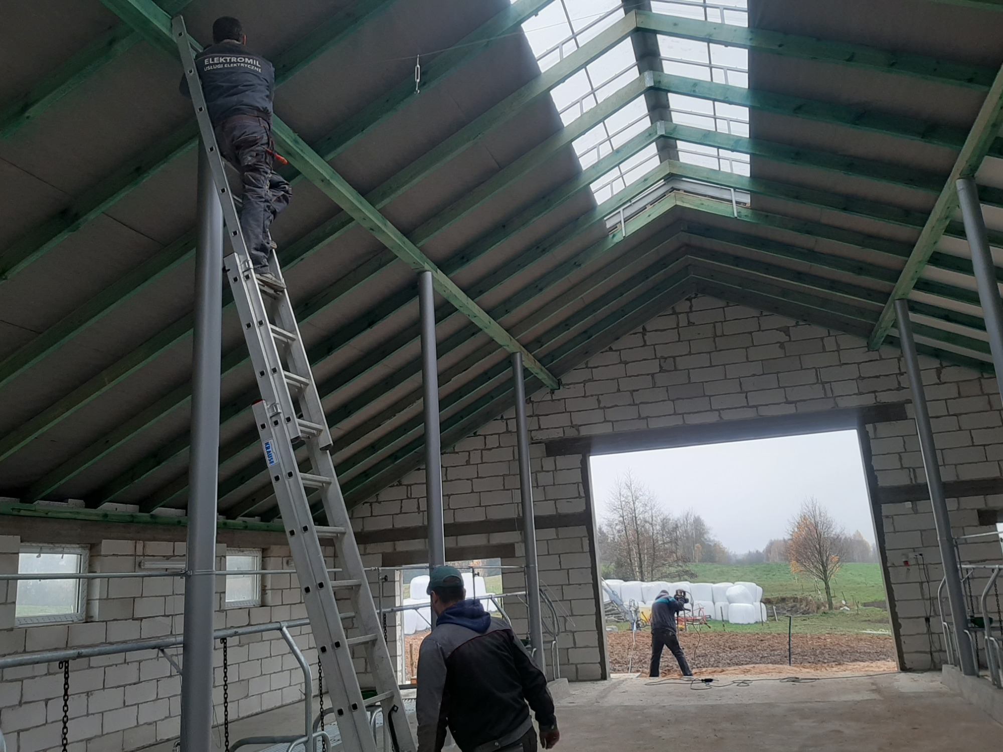 Pracownik z napisem 'ELEKTROMIL USŁUGI ELEKTRYCZNE' na plecach kombinezonu na drabinie wewnątrz budynku gospodarczego z drewnianą konstrukcją dachu i świetlikami, drugi pracownik w tle przy...