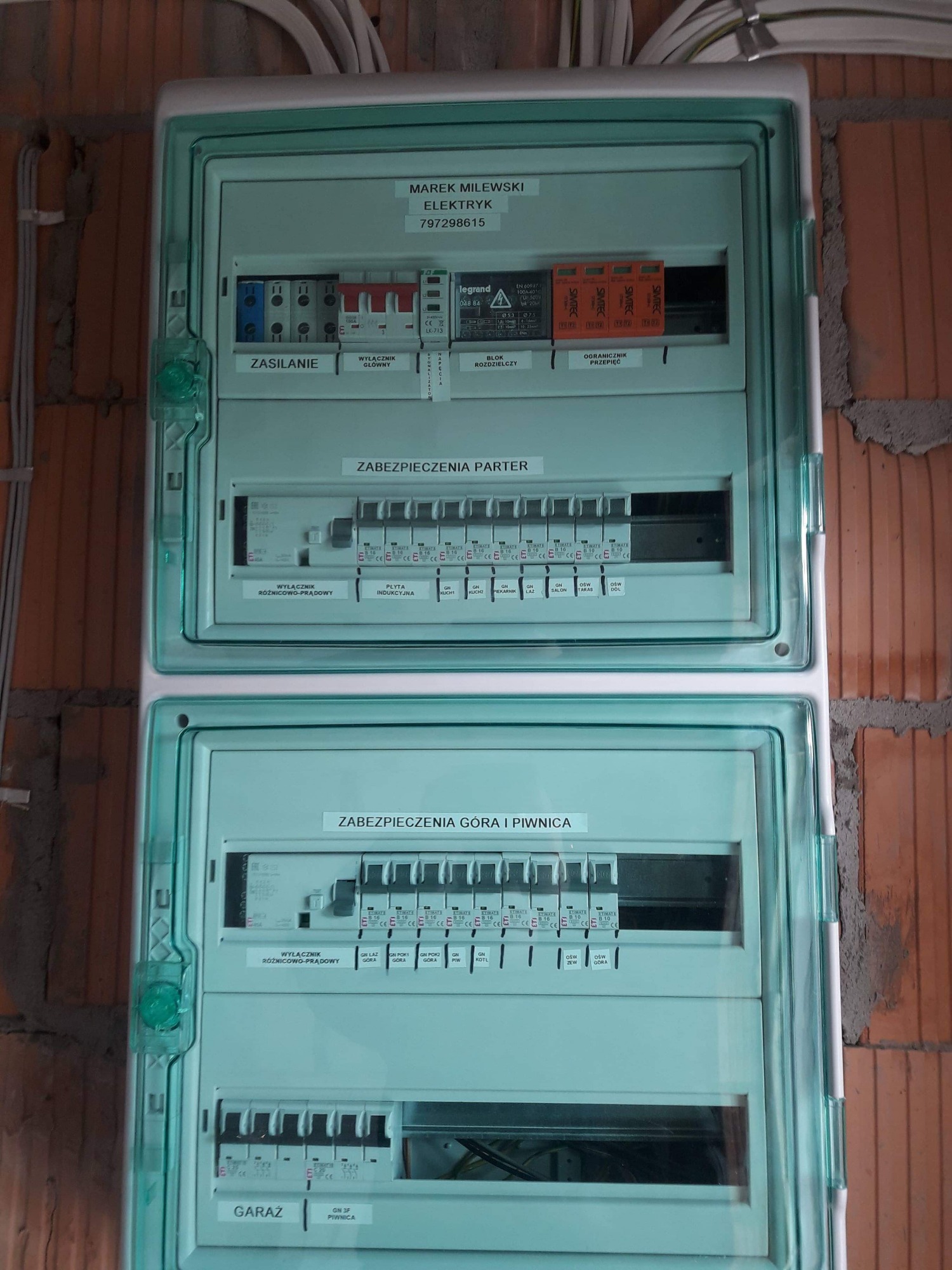 Rozdzielnica elektryczna z bezpiecznikami, opisana etykietami 'Zasilanie', 'Zabezpieczenia parter', 'Zabezpieczenia góra i piwnica', zamontowana na ścianie z cegły, z widocznymi przewodami w górnej...